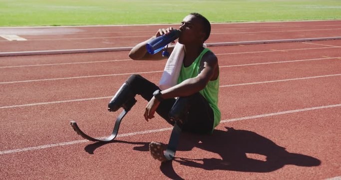 Disabled mixed race man with prosthetic legs sitting on a race track and drinking water