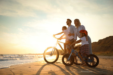 Fototapeta na wymiar Happy parents teaching children to ride bicycles on sandy beach near sea at sunset