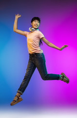 Headphones. Jumping high asian young woman's portrait on gradient studio background in neon. Beautiful female model in casual style. Concept of human emotions, facial expression, youth, sales, ad.