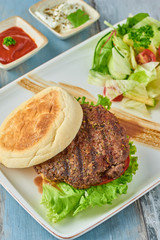 Delicious grilled  burger on white plate on wooden table.  with sauces, french fries and salads.