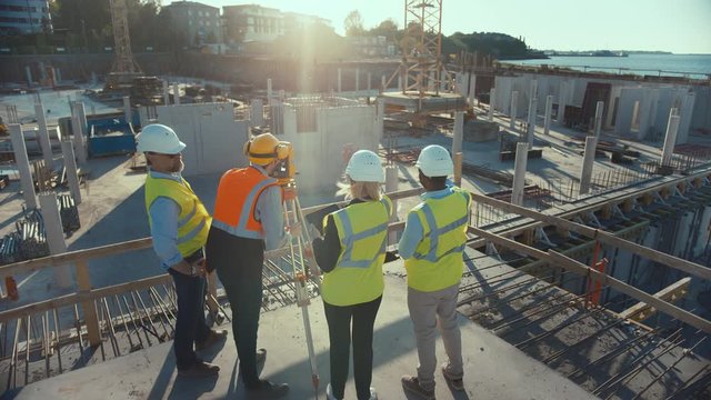 Construction Worker Using Theodolite Surveying Optical Instrument for Measuring Angles in Horizontal and Vertical Planes on Construction Site. Engineers and Architect Discuss Plans Next to Surveyor.