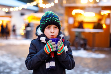 Little cute kid boy drinking hot children punch or chocolate on German Christmas market. Happy child on traditional family market in Germany, Laughing boy in colorful winter clothes