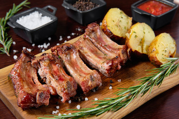 Closeup of pork ribs grilled with BBQ sauce. Tasty snack to beer on a wooden Board for filing on dark wooden background. Nearby are a baked potato and sauces for meat.