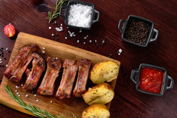 Closeup of pork ribs grilled with BBQ sauce. Tasty snack to beer on a wooden Board for filing on dark wooden background. Nearby are a baked potato and sauces for meat. Top view.