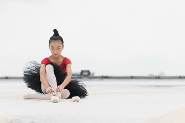 Asian ballerina girl tying pointe shoelace before practicing the ballet dancing sitting on rooftop of the building top view of urban city background.