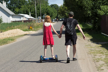 Father and daughter walk along the street of a small town in summer. Child girl rides a hoverboard, sporty dad holds her hand. Back view. Not a staged photo from life.