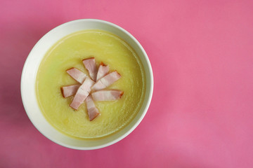 white plate with cream soup of zucchini, potatoes, cream, onions and bacon slices on a pink background top view . cooking at home