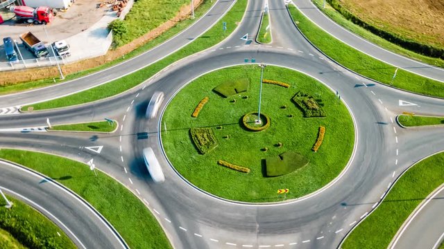 Aerial hyperlapse of roundabout in a residential area with green grass and field. Rush hours, car drive fast