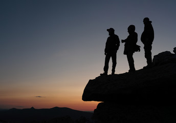 Silhouettes of tourists standing on a cliff against the background of sky after sunset and panorama of the mountains below, panorama.