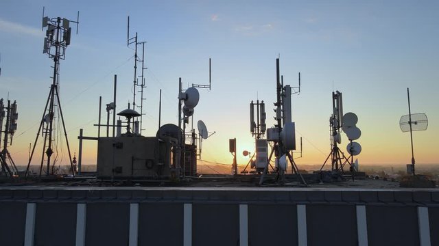 Silhouette of electromagnetic towers with satellite dish, microwaves and panel antennae at rooftop station during golden. Motion footage.