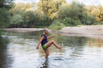 Fototapeta premium Senior woman in a shallow river playing with water. Mature woman enyoing in a river, splashing water with a leg.