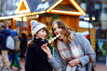 Fototapeta na wymiar Mother and daughter eating white chocolate covered fruits and strawberry on skewer on traditional German Christmas market. Happy girl and woman on traditional family market in Germany during snowy day