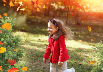 A happy little girl runs in the park with autumn leaves in a bright red coat and gray knitted...
