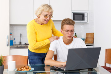 Senior woman hugging her adult son working with laptop at kitchen