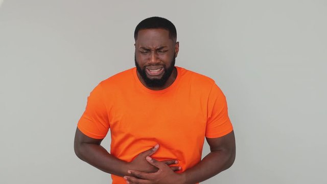 Young sad sick bearded african american man guy male 20s wearing orange basic casual t-shirt look at camera put hands on tummy have stomach ache pain isolated on grey color gray background in studio