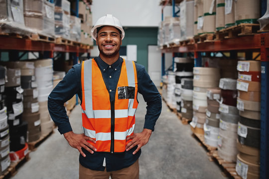 Portrait of successful manager standing in warehouse between shelf filled with goods wearing a white helmet and orange vest for protection with hands on waist - Powered by Adobe