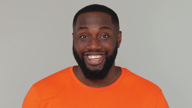 Close up of young bearded african american man guy male 20s in orange basic casual t-shirt looking at camera with charming toothy white-toothed smile isolated on grey color gray background in studio