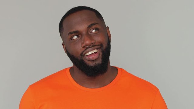 Close up young dreamful bearded african american man guy 20s in orange basic casual t-shirt looking around think dream charming smile blinking wink eye isolated on grey color gray background in studio