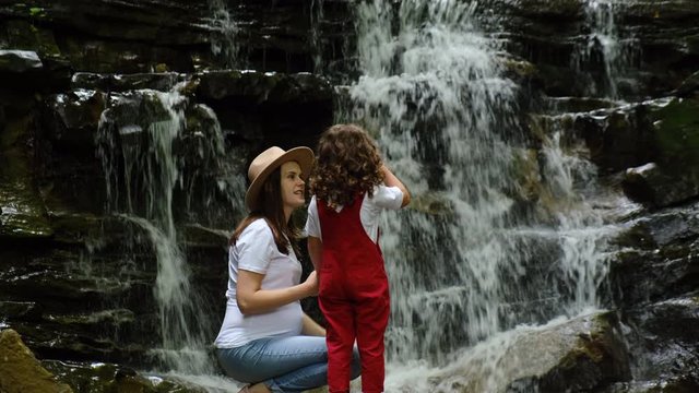 Cheerful affectionate little kid girl kisses happy young mom in hat, embracing affectionate mum having fun standing near mountains waterfall. Concept of family relationships and nature recreation