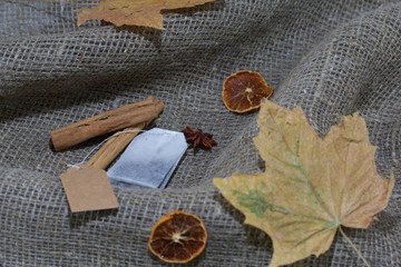 Tea bag, dried maple leaves on a rough linen cloth. Nearby are cinnamon, dried orange slices and anise. Autumn still life.