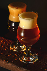 Two tulip beer glasses on dark wooden table over black background. Dark and light beer, stout beer, wheat beer.