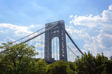 the Manhattan side suspension tower of the George Washington Bridge, a double-decked suspension bridge, above treetops against a bright blue sky with fluffy white clouds with copy space