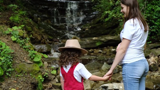 Pregnant mother and preschool daughter in hat stand by mountain waterfall in middle of beautiful forest, enjoying view and breathing fresh air. Outdoor activities travel and family relations concept