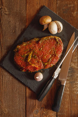 Slice of spiced raw beef with garlic and mushroom on slate plate with meat knife and fork on wooden table