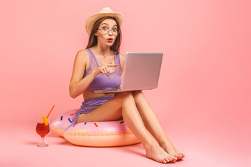 Obraz na płótnie Canvas Funny young woman in swimsuit isolated on pink background. People summer vacation rest concept. Sit in swim inflatable ring, work on laptop pc computer.
