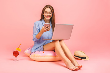 Obraz na płótnie Canvas Funny young woman in swimsuit and casual shirt isolated on pink background. People summer vacation rest concept. Sit in swim inflatable ring, work on laptop pc computer.