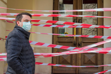 Handsome young European man wearing disposable protective medical face mask, during pandemia of Coronavirus or Covid-19, near closed restaurant or shop door with red protective tape.