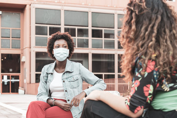 two young women sitting on a bench, chatting using the protective mask and the social distancing, multiracial couple of friends, filter effect vintage