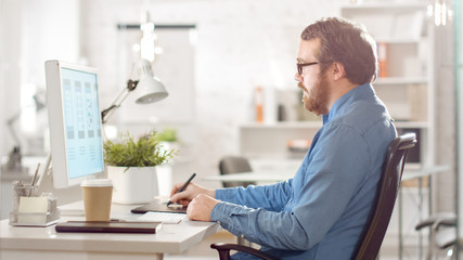 Young Bearded Man Working on His Personal Computer and Taking Notes while Sitting in Bright Modern...