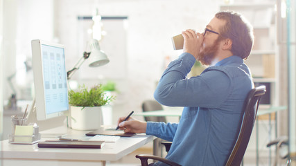 Young Bearded Man Taking Notes while Working on Desktop Computer. He is Drinking Coffee. He Sits in a Creatively Designed Office.