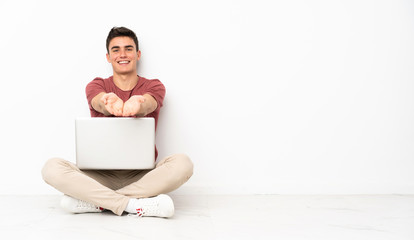 Teenager man sitting on the flor with his laptop holding copyspace imaginary on the palm to insert an ad