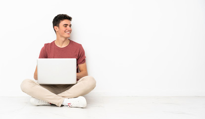 Teenager man sitting on the flor with his laptop with arms crossed and happy