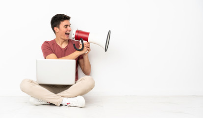 Teenager man sitting on the flor with his laptop shouting through a megaphone