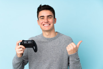 Teenager caucasian man playing with a video game controller isolated on blue background pointing to the side to present a product