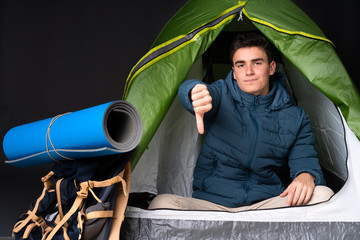 Teenager caucasian man inside a camping green tent isolated on black background showing thumb down...