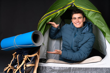 Teenager caucasian man inside a camping green tent isolated on black background holding copyspace to insert an ad