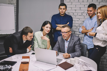 Executive experienced director in eyeglasses with team of creative employees working on creating new advertising for new architectural project sitting at laptop computer and sketches during conference