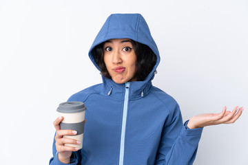 Mixed race woman wearing winter clothes with the hood on and holding a hot takeaway coffee over isolated white background making doubts gesture while lifting the shoulders