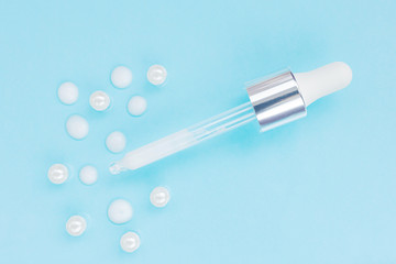 Cosmetic pipette with serum on blue background. Around - drops of serum and pearl. Stylish concept of organic essences, beauty and health products. Copy space, minimalism, flat lay. Modern apothecary.