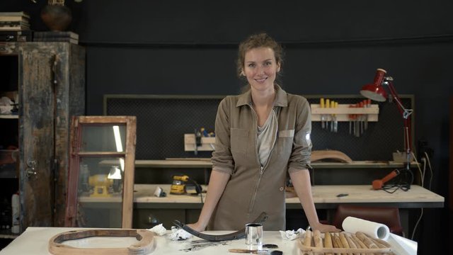 Female restorer smiling at the workshop, standing next to her working table. Gimbal shot of female restoring wooden products, young professional in furniture restoration