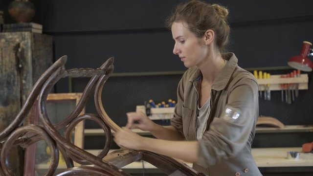 A working female carpenter peels off paint from rocking chair legs with a spatula, restorer in a workshop, handheld shot. Restoration of wooden rocking chair in a workshop. 