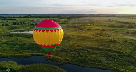 Travel in the air at sunset. Hot air balloon flying in the countryside. Colorful aerostat over the fields and lakes. Aerial view. Motion camera around.