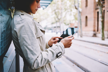 Side view of casual dressed hipster girl choosing application to download on modern touch pad device via free 4G internet connection.Young woman reading e-book standing in urban setting on tram stop