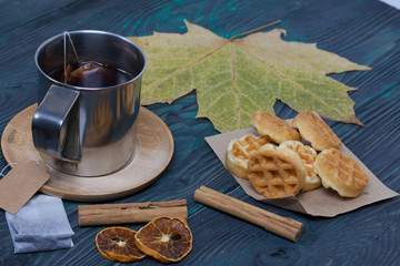 Mug of tea and waffles. Next to it is a tea bag, spices and dried maple leaves. On painted boards. Autumn still life.