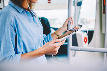 Cropped image of woman in electronic headphones in public transport watching online video during travel trip connecting to 4g wireless for networking, female blogger listening music podcast