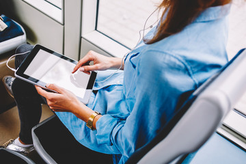 Cropped image of woman sitting inside bus using modern tablet for chatting in application with friends online, female watching video on touchpad with blank screen and in earphones in transport